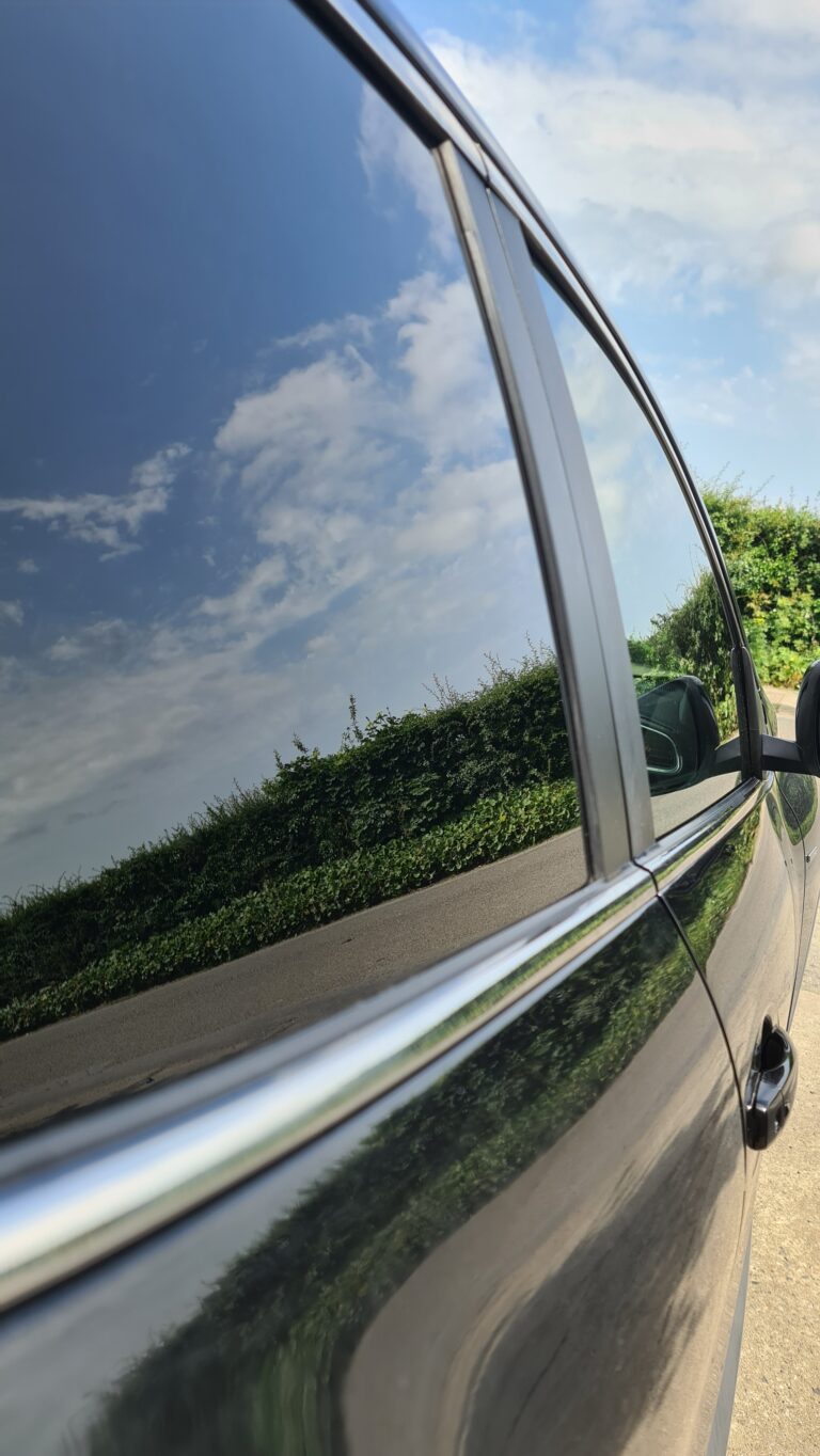 Close-up of a dark saloon's tinted rear door glass reflecting a blue Wexford sky